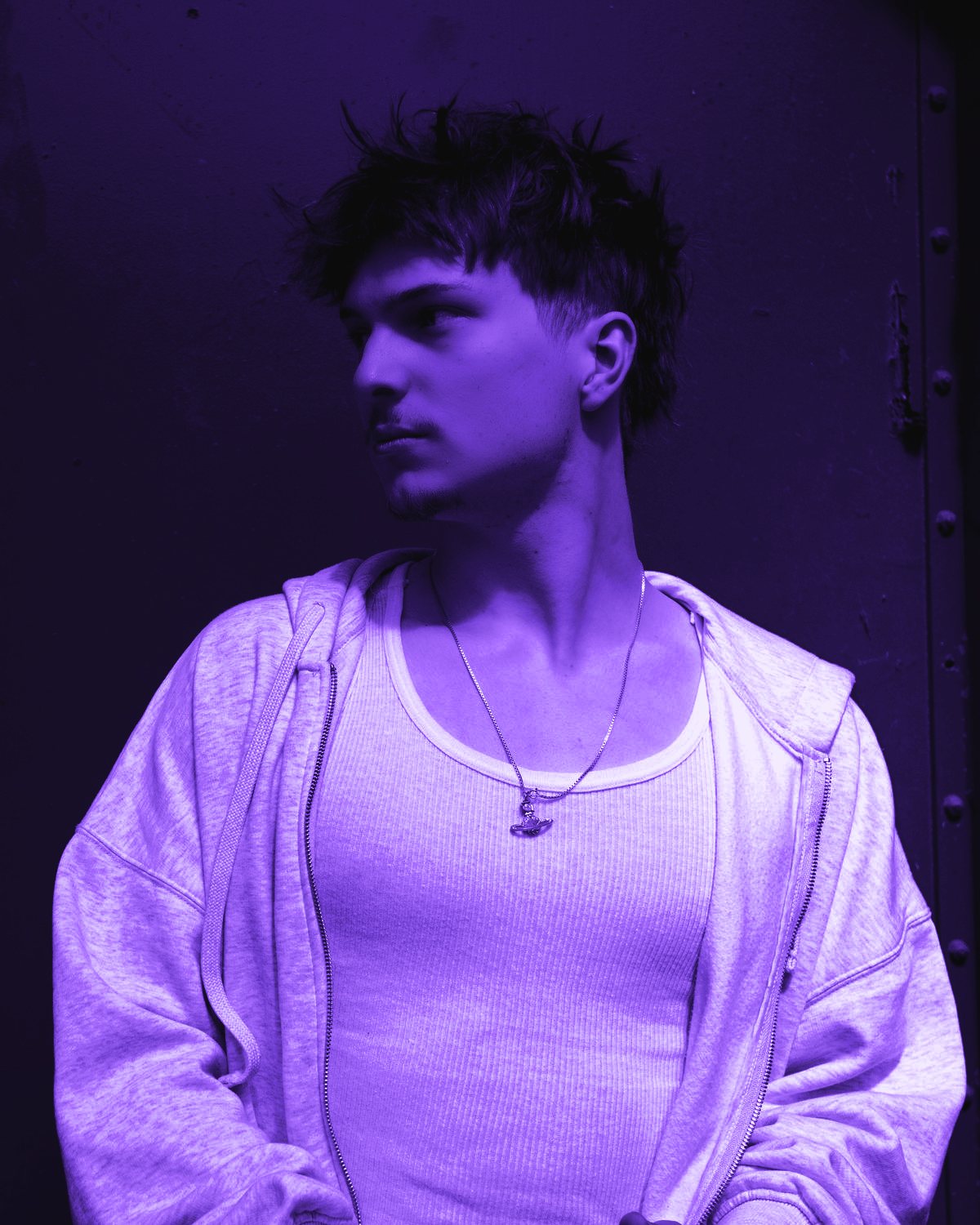 BEJO in a hoodie and tank top, looking to the side against a metal wall, in purple tones